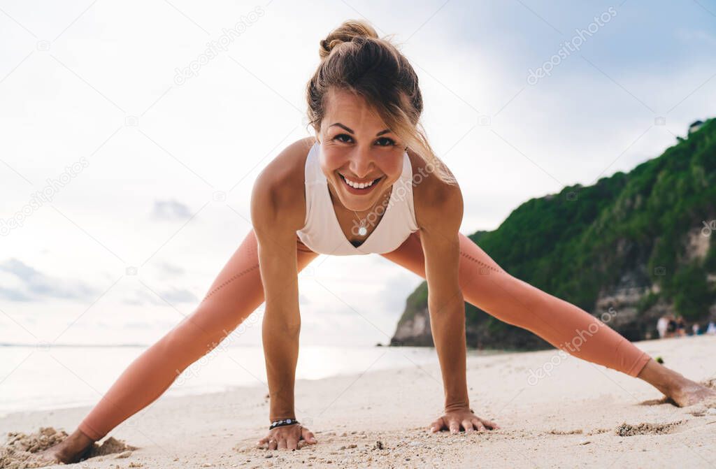 Cheerful woman wearing sportive clothing standing in Wide Legged Forward Bend position during yoga workout on rocky seashore in summertime