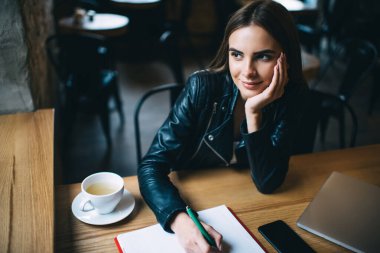Deri ceketli düşünceli bayan öğrenci kafeterya masasında çay fincanı ve eğitim kitabıyla oturuyor ve içerik fikri ve üniversite ödevi, bilgi öğrenme kavramı hakkında düşünüyor.