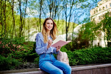 Kot pantolon giyen neşeli genç kız öğrenci şehir parkında beton çitte oturuyor ve kameraya bakarken ajandanın sayfasını çeviriyor.