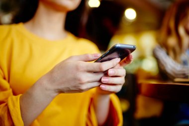 Tanımlanamayan uzun saçlı, sarı bluzlu, uzun kollu, internette gezen, bulanık arka planda cep telefonu kullanan düşük açılı genç bir kadın.