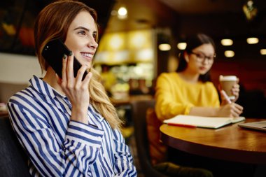 Gülümseyen genç bayan yöneticinin cep telefonuyla konuşması ve iş yerinde bayan meslektaşıyla çalışmasının yan görüntüsü.