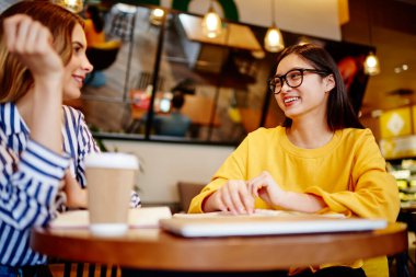 Kafede oturan, kahve içen ve birlikte proje üzerinde çalışırken birbirlerine bakan mutlu çok ırklı kız öğrencilerden.