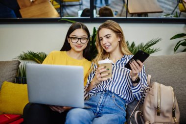 Kafedeki kanepede kahve içerken bilgisayarın ekranına bakıp gülümseyen genç kız öğrenciler.