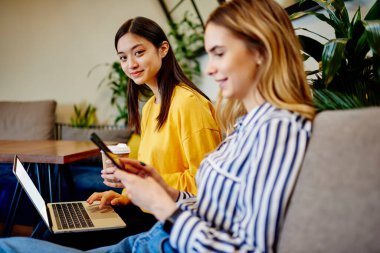 Keyifli Asyalı serbest çalışan, günlük kıyafetleriyle dizüstü bilgisayar kullanan ve gündüz kafede gülümseyen arkadaşının yanında oturan akıllı telefonuyla mesajlaşan.