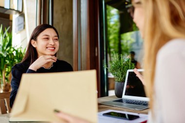 Neşeli Asyalı iş kadını, güpegündüz kafenin terasında iş arkadaşıyla iletişim kurarken elinde cihazlar ve belgelerle masada oturuyor.