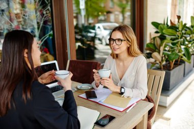 Sırıtkan çok ırklı iş arkadaşları projenin detaylarını anlatırken aynı zamanda not defterleri ve aletlerle masaya oturuyorlar ve gündüz vakti kafenin terasında kahve içiyorlar.