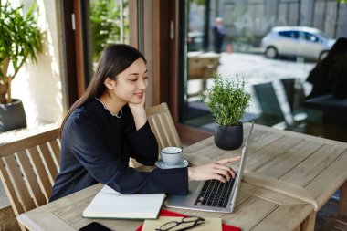 Genç Asyalı serbest çalışan yüksek açılı bir bayan dizüstü bilgisayarı ve sıcak bir kahve bardağıyla ahşap masada oturuyor.