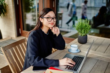 Asyalı bir kadın gündüz vakti elinde içki ve verandada telefonla oturup kameraya bakarken bilgisayarla uzaktan kumandayla çalışıyor.
