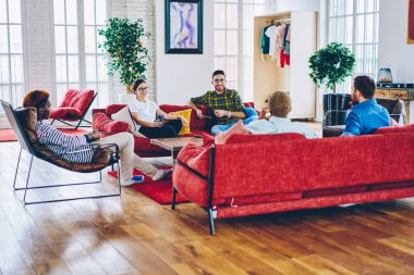 Modern apartmandaki rahat kırmızı koltukta otururken rahat bir şekilde sohbet eden en iyi arkadaşlar. Çatı katının iç dekorasyonunda çeşitli hipsterler birlikte boş zaman geçiriyorlar.