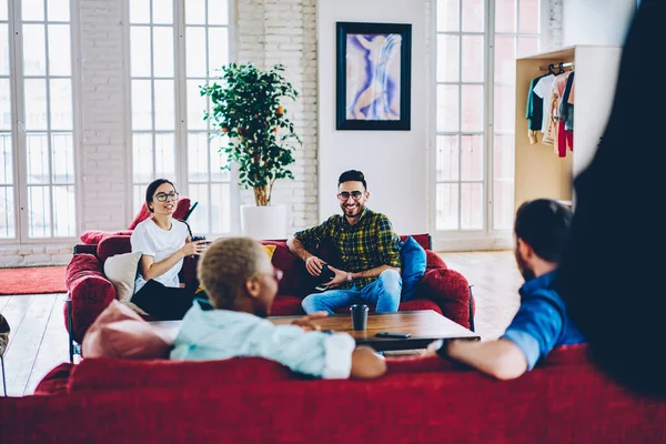 Görsel koruma için optik gözlüklü neşeli Türk adam elinde kahve fincanı ve kırmızı kanepeli çatı katındaki dostça görüşmede gülümsüyor ve hafta sonu sosyalleşmek için toplanıyor.