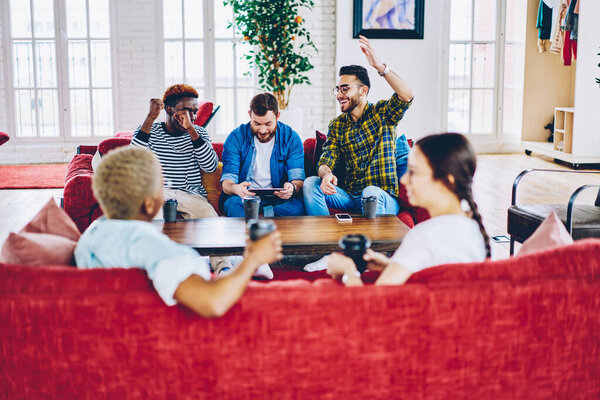 Happy hipster guys feeling satisfied while celebrating victory watching live stream on digital tablet using 4G internet while blurred women talking during home friendly meeting in modern apartment