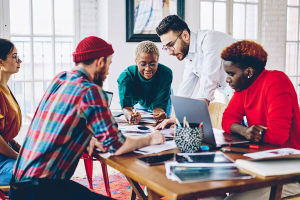 Multicultural creative coworkers casual wear discussing ideas and freelance work during together briefing at meeting table with laptop, young group of programmers collaborating and talking indoors