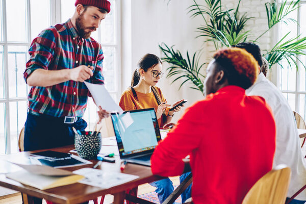 Caucasian man with paper report talking about business strategy while cooperating with diverse colleagues, professional hipster guy communicate with multiracial partners about graphic design