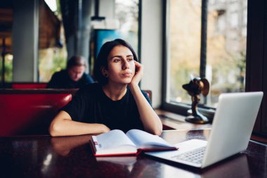 İş sırasında dizüstü bilgisayar ve planlayıcıyla masaya yaslanmış bir şekilde otururken günlük giysiler içinde sakin bir etnik kadın uzaktan çalışan.