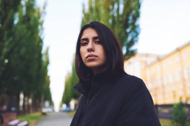 Düşük açılı, siyah saçlı, düşünceli, etnik bir kadın. Arka planda bulanık bir şekilde şehir caddesinde kameraya bakıyor.