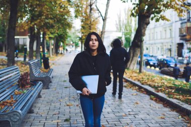 Sokakta yürürken ve dizüstü bilgisayar taşırken el ele kameraya bakan, gündelik kıyafetli, ciddi bir etnik kadın.