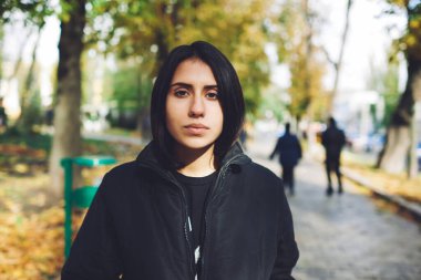 Siyah elbiseli genç, düşünceli, siyah saçlı, güneşli bir sonbahar gününde caddede yürüyen kameraya bakan bir kadın.