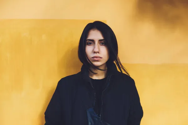 昼間通りに黄色の壁の横に立ってカメラを見て長い黒い髪の黒いジャケットを持つ若い民族の女性 — ストック写真