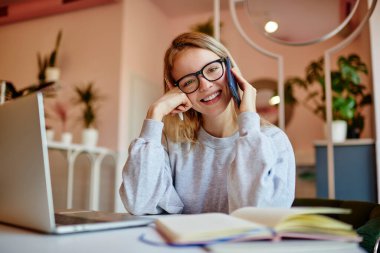 Optik gözlüklü neşeli bayan öğrencinin portresi cep telefonunu kullanarak birlikte çalışmada olumlu bir konuşma yapıyor, modern dizüstü bilgisayarlı mutlu bir kadın akıllı telefon uygulaması aracılığıyla arıyor.