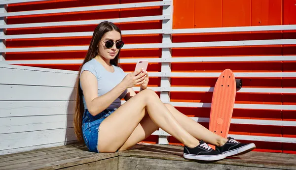 千年女性 戴着时髦太阳镜 在室外休息 与互联网连接 阅读有关极端爱好的出版物 带着铜板的滑板 用智能手机装置在网上聊天 — 图库照片