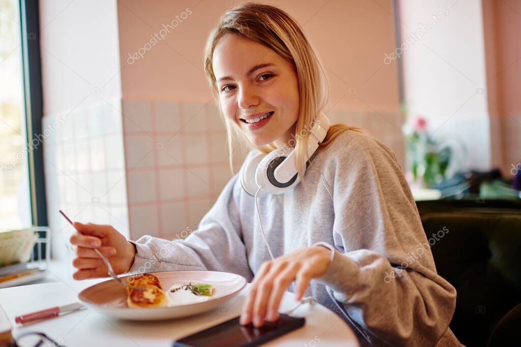Portrait of happy Caucasian woman with electronic headphones and modern smartphone device enjoying lunch time in cafeteria, cheerful hipster girl with mobile phone posing during cafe breakfast