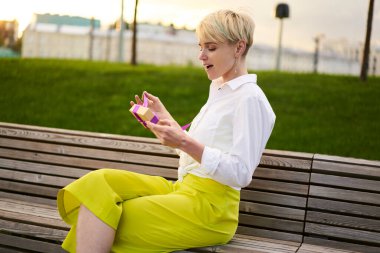 Moda giyinmiş 20 'li yaşlarda kadınlar ahşap bankta dinleniyor açık hava kutusunu açarken hediye alıyorlar, paketini açan beyaz yeniyetme kız hediye paketini açarken şok olmuş hissediyorlar.