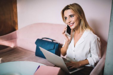 Genç bayan girişimci resmi giysiler içinde masada dizüstü bilgisayarla oturuyor ve modern çalışma alanında proje üzerinde çalışırken telefonla konuşuyor.