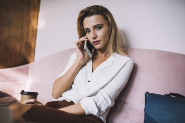 Düşünceli bir kadın, şık gündelik kıyafetlerle kameraya bakarken yumuşak pembe koltukta kahve alıp telefonla konuşuyor.