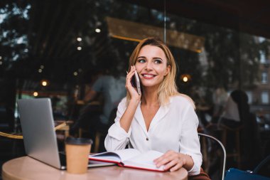 Şık giyinen neşeli kadın girişimci netbook ile masada oturuyor ve cep telefonunu açıyor ve gündüzleri kafenin terasında çalışırken konuşuyor.