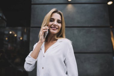 Uzun saçları boyalı, zarif bluzunu giymiş, dışarıda kameraya bakıp iş arkadaşının telefon konuşmasını dinleyen genç bir hanım.