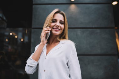 Uzun saçları boyalı, zarif bluzunu giymiş, dışarıda kameraya bakıp iş arkadaşının telefon konuşmasını dinleyen genç bir hanım.