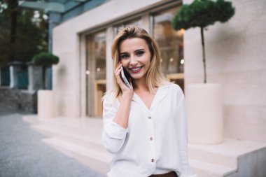 Gündüz vakti modern binanın girişinde dikilirken beyaz bluzlu neşeli sarışın kadın akıllı telefonlara cevap veriyor.
