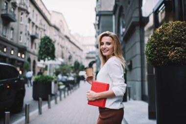 Tarz sahibi beyaz bluz ve kahverengi etekli neşeli genç bayan girişimci kaldırımda duruyor ve kameraya bakıp kahve içiyor.