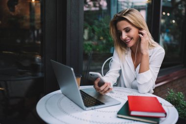 Kulaklık takan, cep telefonlu olumlu bir kadın açık kafede çalışırken elinde netbook ve defterlerle masada oturuyor.