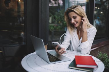 Kulaklık takan, cep telefonlu olumlu bir kadın açık kafede çalışırken elinde netbook ve defterlerle masada oturuyor.