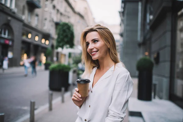 白いブラウスを着た幸せな若い女性 街の店の入り口近くの歩道に立ってコーヒーの使い捨てカップと離れて見て — ストック写真