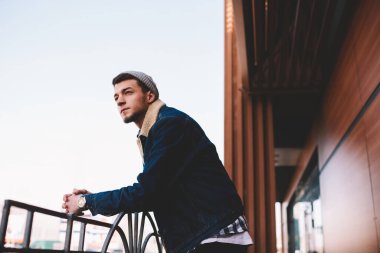 Şık giyinmiş yakışıklı bir erkek sokakta dikilip ahşap evin yanındaki korkuluklara yaslanmış duruyor.