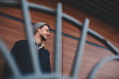 Şapkalı, dişlek gülüşlü, sıradan giyinmiş genç bir erkek ahşap duvarlı binanın yanında dikiliyor.