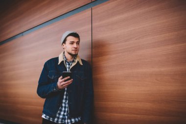 Odaklanmış, gri şapkalı genç bir erkek. Binanın yanında duruyor, ahşap duvarlı ve cep telefonlu sörf yapıyor.