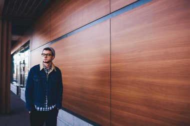 Genç sakallı bir erkek, gözlüklü ve elleri ceplerinde, modern binanın yanında ahşap duvarla yürürken başka tarafa bakıyor.