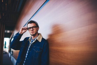 Ciddi sakallı, sıradan giysiler içinde, sokağın ahşap duvarına yaslanmış ve gözlük çerçevesine dokunmuş bir şekilde başka tarafa bakan bir erkek.