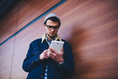 Ahşap duvarın yanında duran ve sokakta mesajları kontrol etmek için tablet kullanan sıradan giysili dalgın bir erkek.