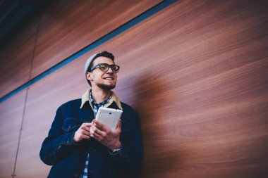 Kalın elbiseli, şapkalı ve dişlek gülüşlü yakışıklı bir erkek, tahta duvarın yanındaki sokakta tablet kullanırken başka tarafa bakıyor.