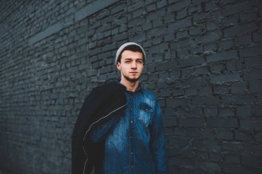 Kot gömlekli, gri şapkalı, omzunda koyu renk ceketli, sokakta yüksek koyu tuğlalı duvar boyunca yürüyen ciddi bir etnik erkek.