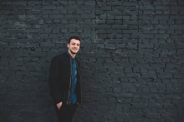 Koyu gri tuğla duvarın yanında duran kot gömlekli ve siyah ceketli yakışıklı hippi erkek kameraya bakıyor.