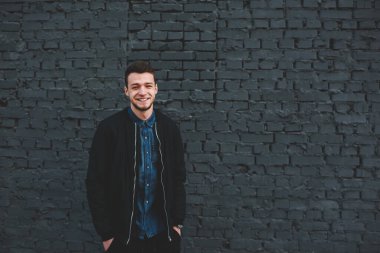 Neşeli, etnik sakallı, sıradan giyinmiş ellerini cebinde tutan ve gülümseyen genç bir erkek sokakta tuğla duvarın yanındaki kameraya bakıyor.