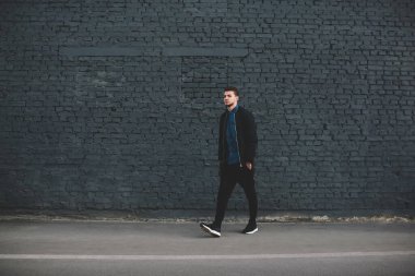 Moda kıyafetleri içinde, sokakta yüksek gri tuğla duvar boyunca yürüyen düşünceli, etnik sakallı genç bir adam.