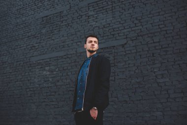 Kot gömlekli, siyah ceketli, elini cebinde tutan ve caddedeki yüksek tuğlalı duvarın yanına bakan dalgın genç bir erkek.