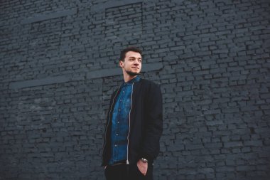 Olumlu genç etnik sakallı erkek, sıradan kıyafetler giymiş, elleri ceplerinde, sokakta tuğla duvarın yanında duruyor ve başka tarafa bakıyor.