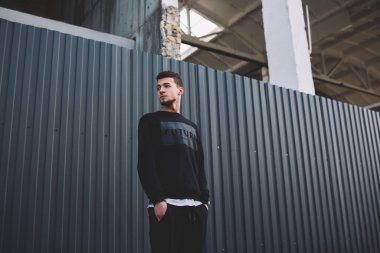 Modaya uygun giysiler içinde, elleri ceplerinde metal çitlerin arkasına bakan, düşük açılı, etnik, ince sakallı bir erkek.